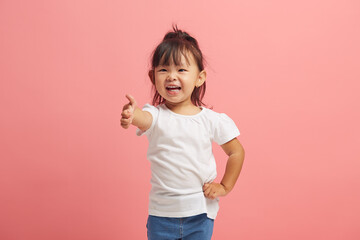 Cheerful and happy little asian girl smiles and shows thumb up gesture standing over pink isolated...