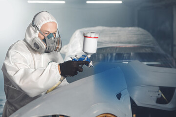 Car paint worker spraying blue paint to car body element using spray gun in vehicle workshop chamber. Complex restoration of automobile body. 
