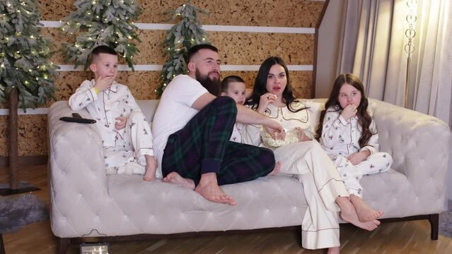 Cheerful mom, dad and three little kids in pyjamas eating popcorn on sofa while watching family movie at cozy decorated xmas home. Winter holidays, new year 2025 concept. Family day. Happy childhood.