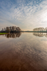 Landscape photo with a large lake and bare trees as silhouettes on the shore. It is a windless day and the water surface is very smooth. The low sun is just breaking through the cloudy sky.