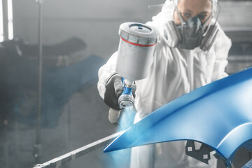 Car paint worker spraying blue paint to car body element using spray gun in vehicle workshop...