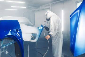 Male Industrial worker sprays paint using airbrush gun on part of car wears in protective mask and...