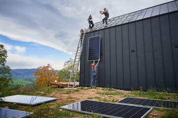 Men roofers installing solar panel system on roof of house. Workers in helmets lifting up...