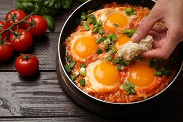 Woman dipping piece of bread into delicious Shakshuka at wooden table, closeup