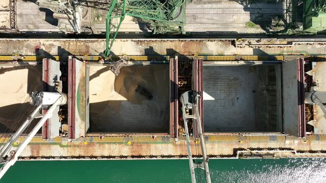 Unloading dry cargo ship by cranes in port. Aerial view of buldozer working in cargo hold