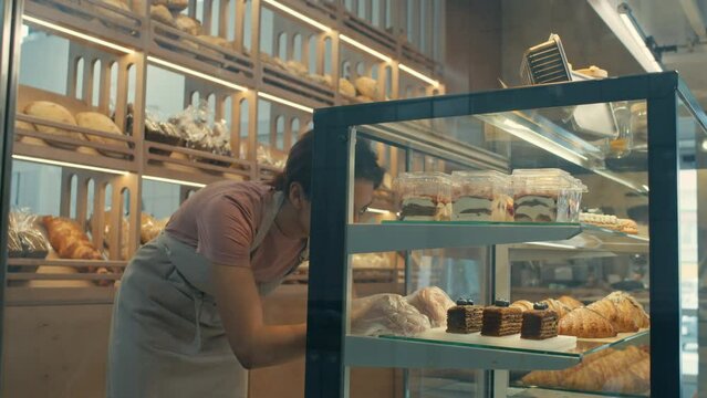 Tilt down shot of female worker in disposable gloves putting desserts on display shelf in bakery