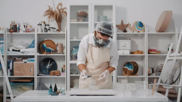 Female artist pouring paint into cup with art epoxy resin working in art studio workshop, creative woman in protective face mask engaged in trendy hobby creating beautiful sea waves acrylic paintings