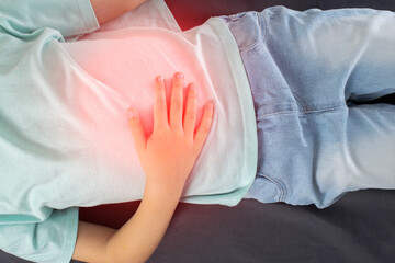 A seven-year-old girl in a green T-shirt lies and holds her sore stomach with her hands. Concept of appendicitis in children, inflammation in the epigastrium. Vomiting and nausea.