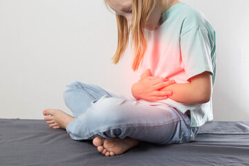 A seven-year-old blonde girl sits and holds her red belly. Concept of pain and cramps in the...
