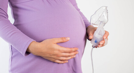 A pregnant girl is holding a nebulizer against the background of her belly. Koznept treatment of...