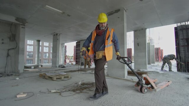 Long slowmo shot of hard-working mid-aged Caucasian construction worker in yellow hardhat carrying heavy hand pallet trolley along concrete floor at unfinished construction site