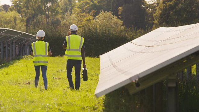Rear view of male and female engineers wearing hard hats and hi vis safety vests walking through field outdoors inspecting solar panels on sustainable energy farm - shot in slow motion