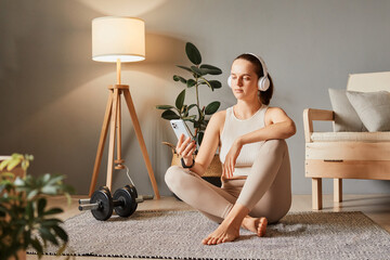 Online health and fitness. Mobile phone in fitness. Break time with technology. Exercising with a cell phone. woman wearing beige tracksuit sitting on floor using gadget - obrazy, fototapety, plakaty