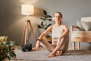 Fotobehang Active care for body. Leisure in a sporty lifestyle. Sport and fitness harmony. Positive woman wearing beige sportswear relaxing after home work out sitting on floor in living room looking at camera © sementsova321