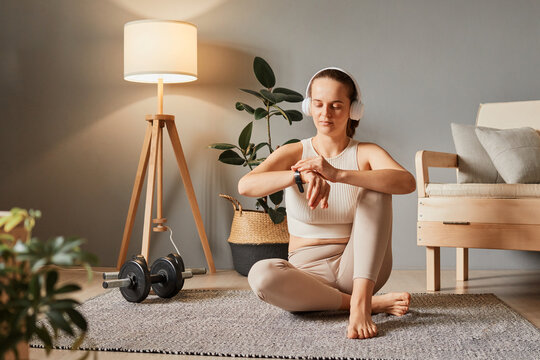 Digital wellbeing at home. Smart bracelet in sport. Sporty smartwatch features. Sportswoman's tracker. Sporty beautiful woman in headphones checking her workout time at home