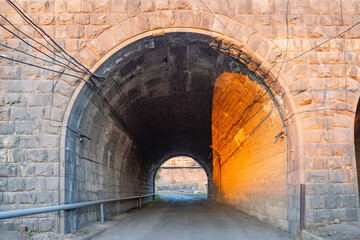 Road tunnel. Road for transport under rock or wall. Stone tunnel in sunny weather. Street tunnel for cars. Asphalt road leads to arches. Path for transportation. Short corridor through rock