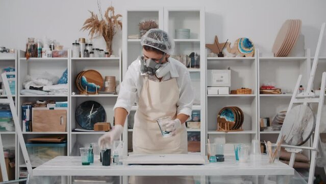 Female artist mixing colored art epoxy resin in plastic cup working in art studio workshop, creative woman in protective mask engaged in trendy hobby creating beautiful sea waves acrylic paintings