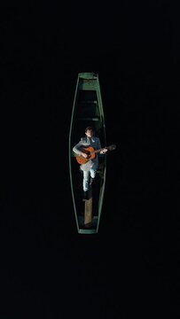 Vertical video. A man in a gray coat lies in a wooden green authentic boat, plays the guitar and sings. Clear dark water surrounds the boatman. Aerial view.