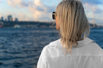 Photo from the back of a young beautiful blonde woman in a white shirt looking at the Bosphorus and...