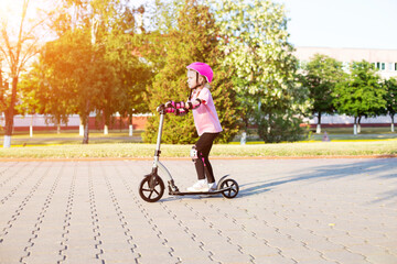 A beautiful girl of 7 years old rides a scooter in a pink protection against injuries and falls. Sunny summer day. Copy space for text