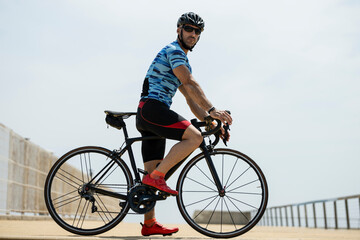 Side view of a fit man standing on bike wearing helmet and sunglasses. Athletic male cyclist training with cycle outdside looking at camera. - obrazy, fototapety, plakaty