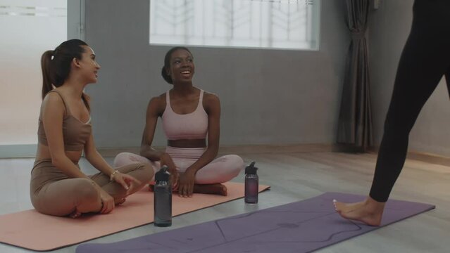 Medium long shot of woman joining others sitting on floor while waiting for stretching class beginning and talking at fitness studio