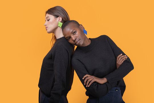 Side view of diverse young female friends leaning on each other looking at camera while wearing trendy colorful earrings against yellow background