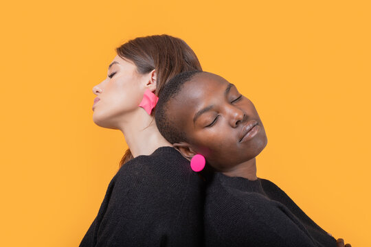 Side view of diverse young female friends leaning on each other with eyes closed while wearing trendy pink earrings against yellow background