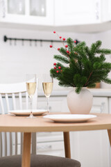 Two glasses of Champagne, two white plates on round wooden table. Christmas home interior. Ceramic vase with fir branches.