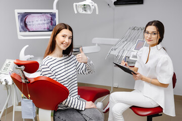 A cute girl is sitting in a dental chair in the dentist's office, smiling and pointing her finger...