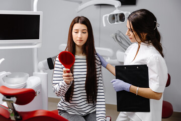 Young pretty girl looking sadly in the mirror while sitting in the dental chair, and the female...
