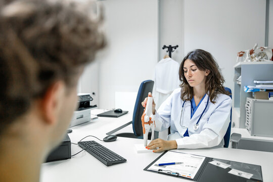 Woman doctor sitting at table with tendon and muscle figurine in clinic and showing to unrecognizable man patient
