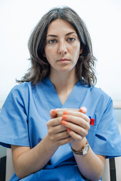 Young female doctor holding cup of coffee and looking at camera while sitting