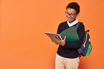 happy african american schoolboy in uniform holding backpack and looking at textbook on orange