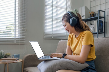 Asian woman sitting on sofa in living room using laptop and listening to music on headphones.