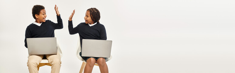 happy african american schoolkids in uniform using laptops and giving high five on grey, banner - obrazy, fototapety, plakaty