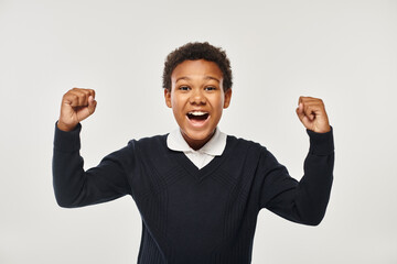 excited african american schoolboy in uniform rejoicing while looking at camera on grey backdrop