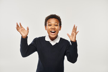 excited african american boy in school uniform rejoicing while looking at camera on grey backdrop