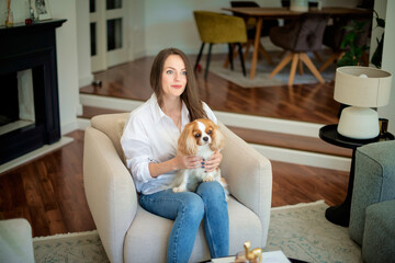 Portrait of an attractive brunette haired woman with her cute puppy sitting in an armchair in her modern home