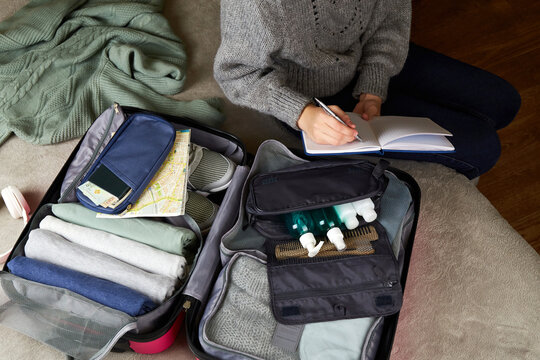 The girl writes down a list of things for the trip in her notebook. An open suitcase with things and accessories for travel.