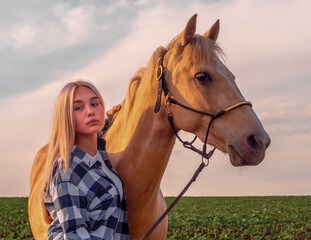 Obraz premium young blonde girl with a beige horse on the ranch on the background of a green field and sky