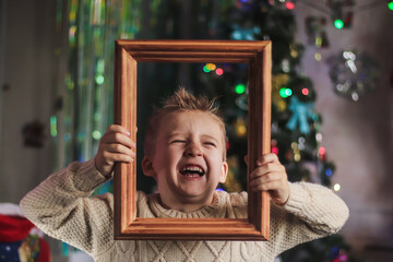 portrait of a cute boy of European appearance in a wooden picture frame against the backdrop of a Christmas tree in the room - obrazy, fototapety, plakaty
