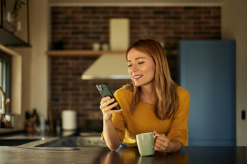 A blonde woman using a mobile phone and holding a cup of coffee in the morning, at home.