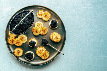Blinis with black caviar and cream cheese, overhead flat lay shot on a festive dish, mini pancakes,...