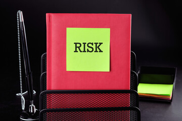 RISK Text on a yellow sticker on a red vertical notepad on a black background next to a pen and...