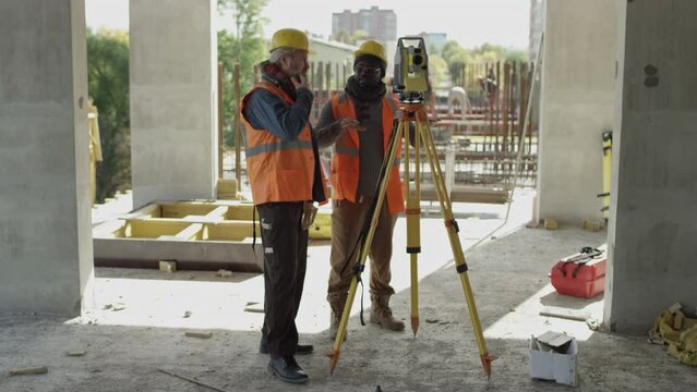 Long full shot of two diverse engineers in hardhats and reflective safety vests standing by total station for angle measuring and having discussion, working together at concrete construction site