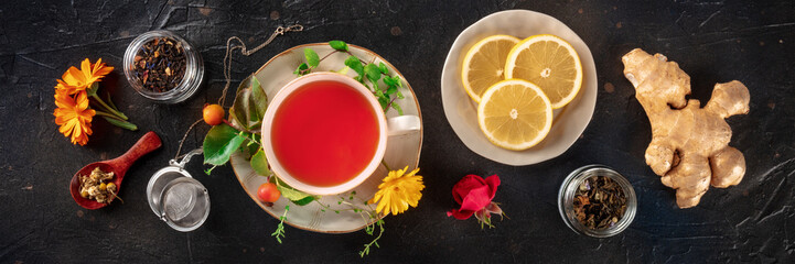 Tea with herbs, fruits, and flowers panorama, overhead flat lay shot on a black background. Healthy...