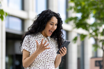 Young joyful woman winner received online notification on phone, Hispanic woman with curly hair...