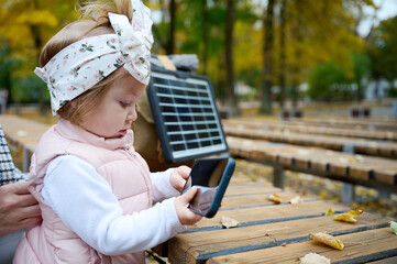 Young cute girl with near bench in the park with fallen leaves, charging phone with portable solar...