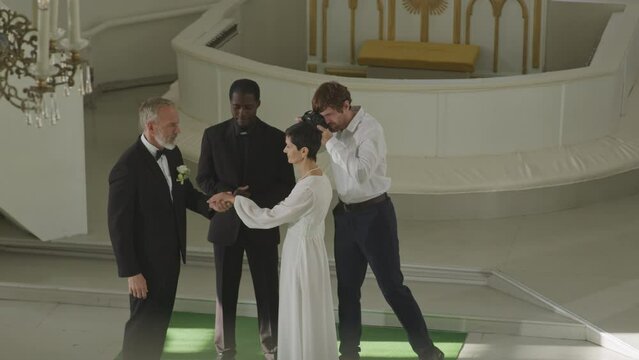High angle slowmo of mid-aged Caucasian man and woman in love exchanging rings, Black male officiant looking at them and smiling and photographer taking pictures of beautiful church wedding ceremony
